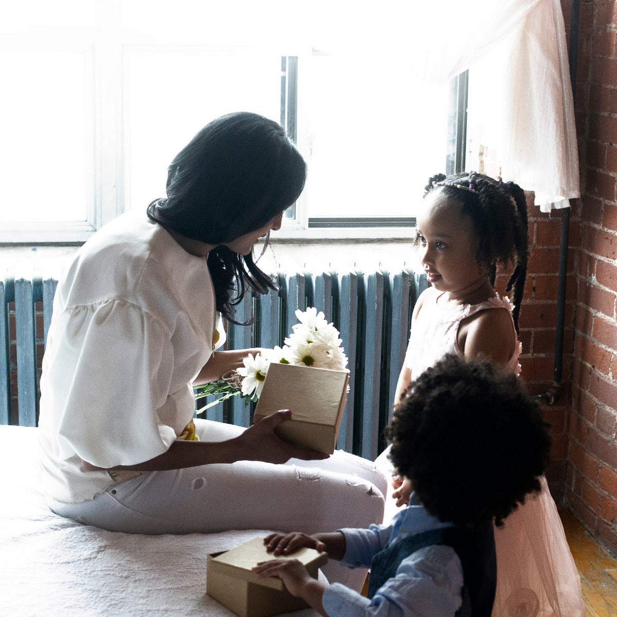 Woman sitting on a bed with two children, holding flowers and a box, in a room with a brick wall and window.