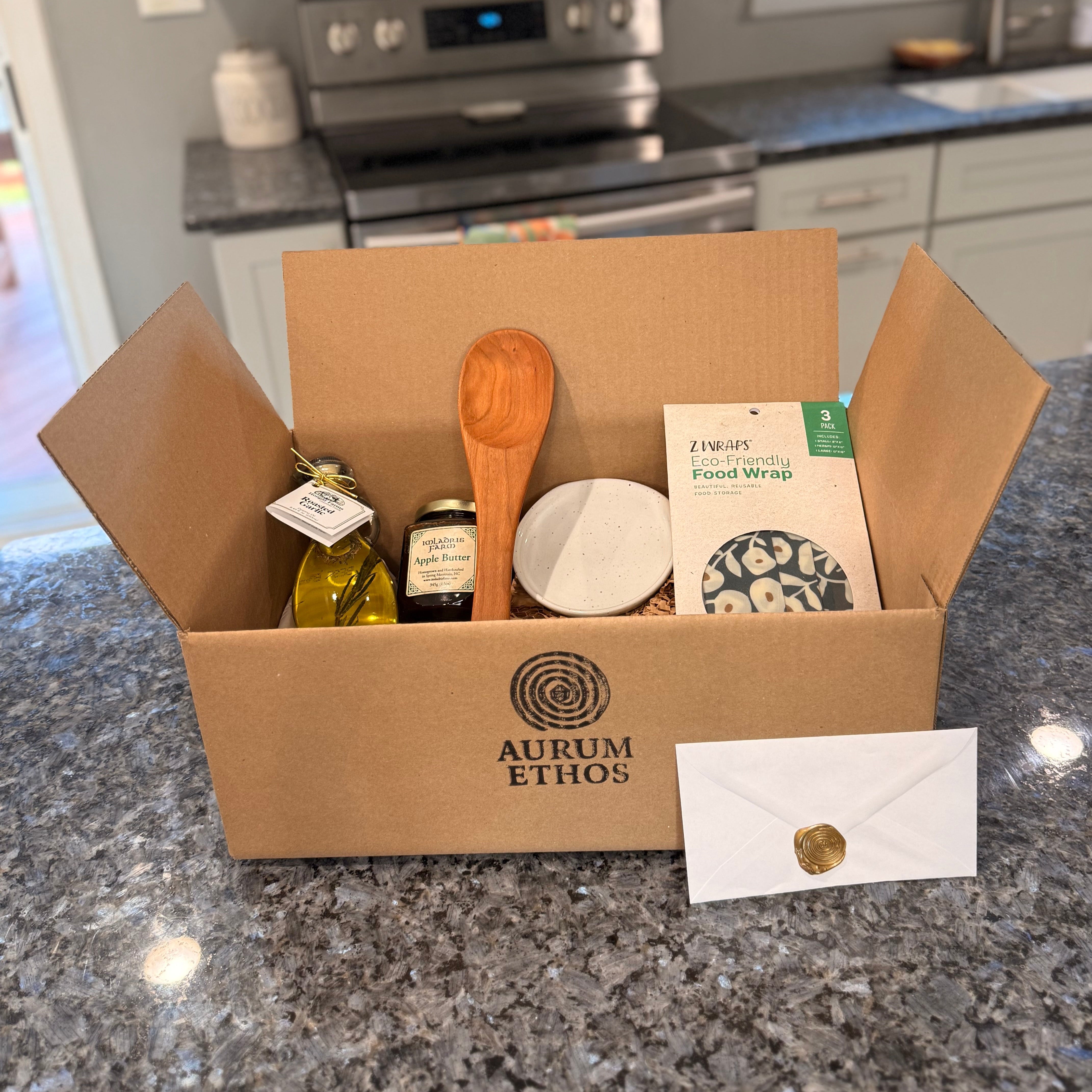 Kitchen Essentials Plus Story Box with a wooden spoon, eco friendly reusable food wraps, a bottle of roasted garlic olive oil, a handmade ceramic spoon rest, and a jar of apple butter.